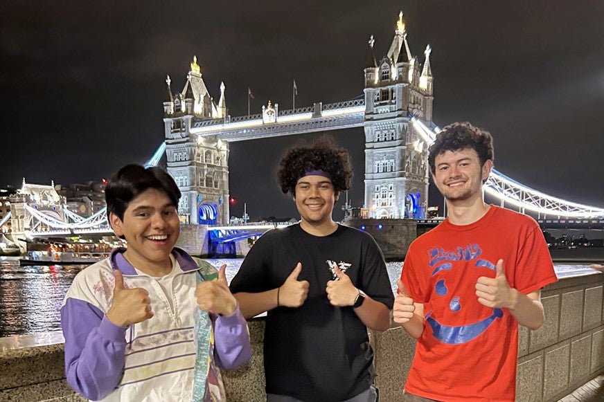 Three diverse young men giving thumbs-up in front of London Bridge.